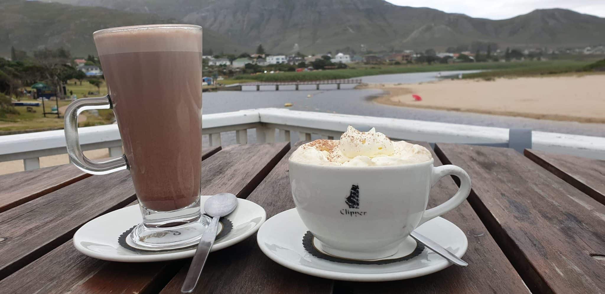 A tall glass of frothy hot chocolate and a cup of whipped cream topped coffee sit on a wooden outdoor table overlooking a river, sandy shore, and distant mountains on a sunny day in Kleinmond.